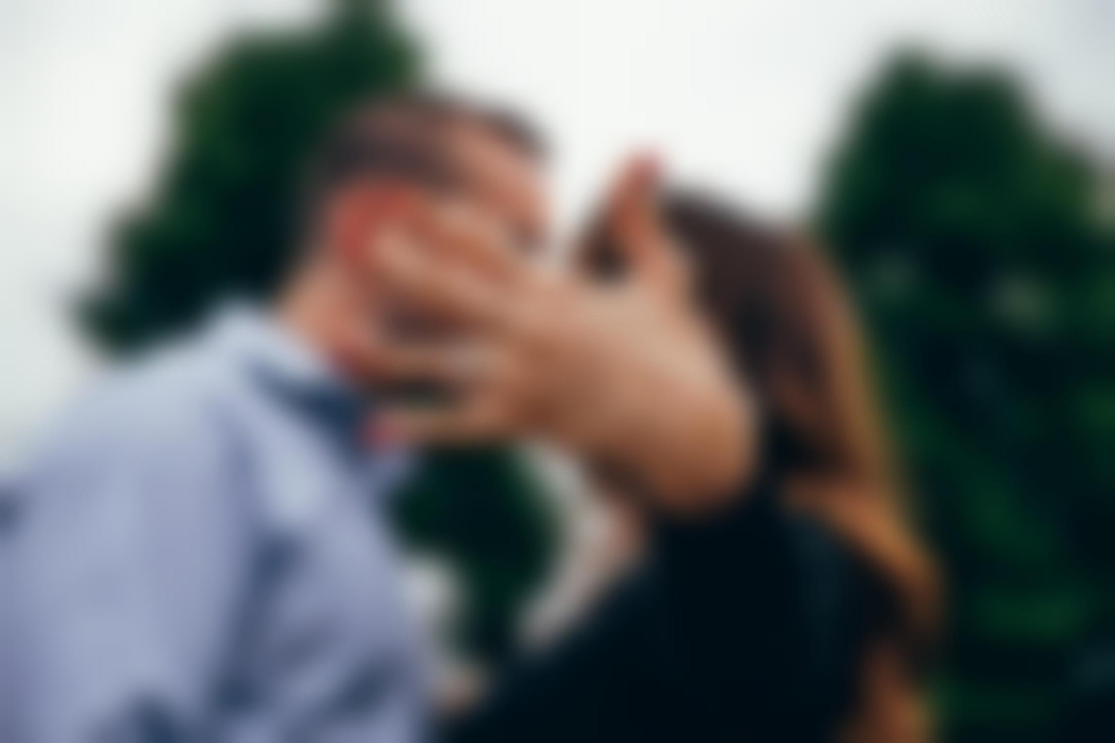 Woman’s hand featuring diamond engagement ring with couple kissing in background.