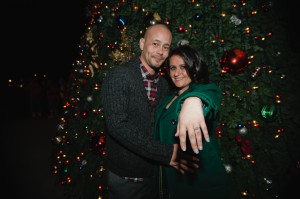 Couple embracing in front of Christmas tree showing of woman’s diamond engagement ring.