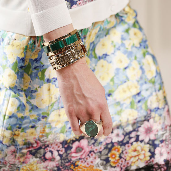 Woman in floral skirt wearing stacked cuff bracelets and a statement ring all set with green gemstones.