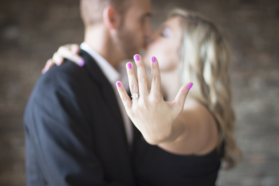 Woman’s hand featuring diamond engagement ring with couple kissing in background.