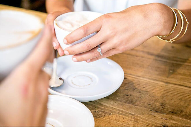 Woman’s hand holding coffee cup featuring yellow gold bangles and white gold diamond engagement ring.