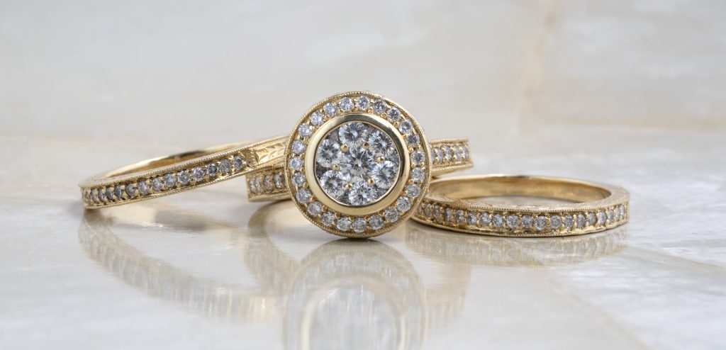 Two wedding bands and one engagement ring all in yellow gold and set with diamonds stacked on a white table.