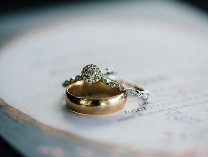 Two diamond engagement rings stacked on a yellow gold wedding band sitting on a white table.