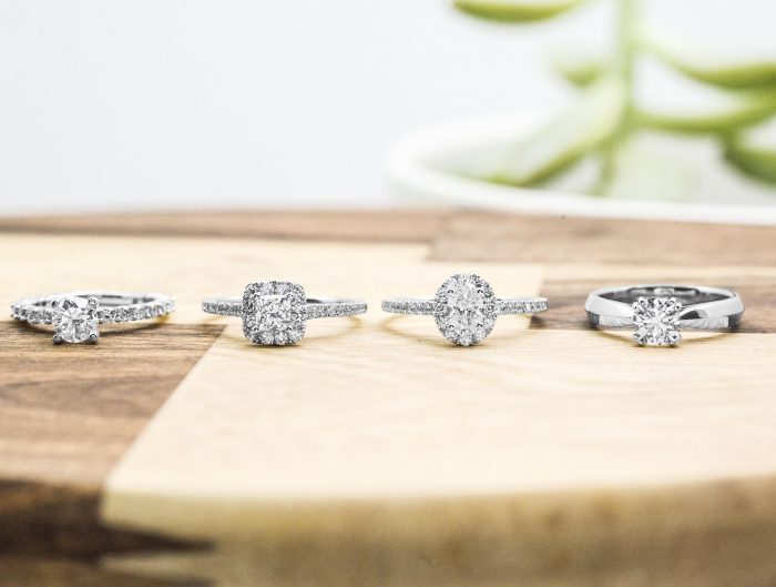 Four white gold diamond engagement rings of various shapes lined up on a wooden table.