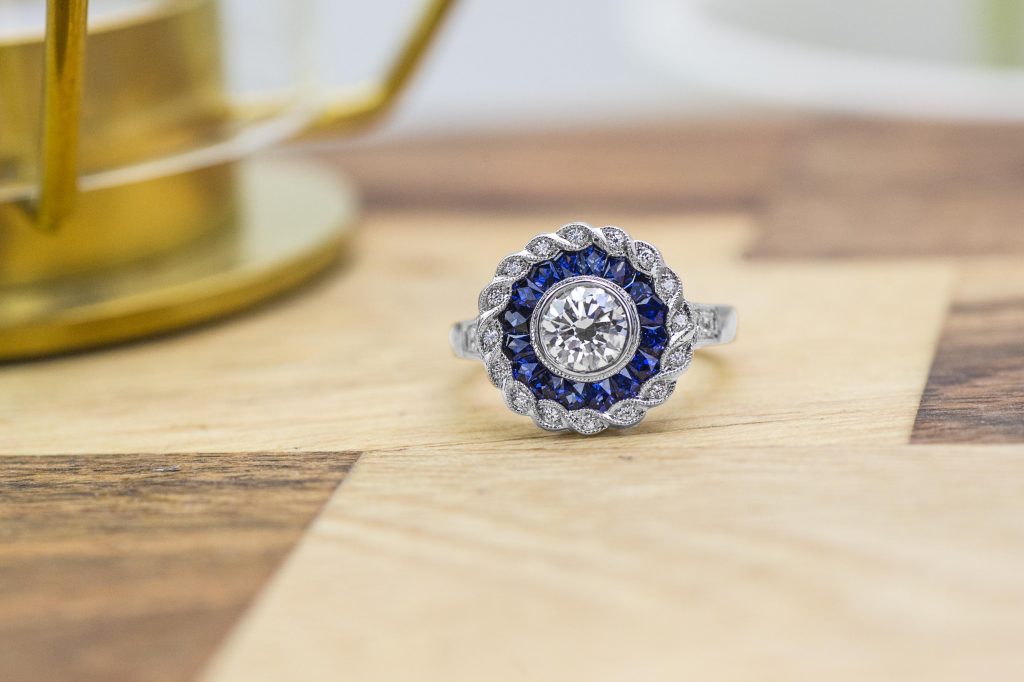 White gold engagement ring centered with a diamond surrounded by a double halo of
blue sapphires and diamonds with diamonds in the band.