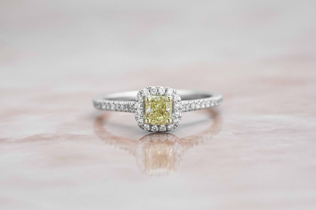 White gold engagement ring centered with a light fancy yellow diamond surrounded by a white diamond halo and white diamonds in the band.