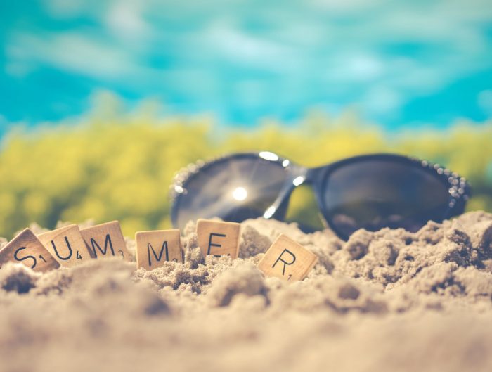 Black sunglasses in the sand at the beach with Scrabble tiles spelling out “Summer”.