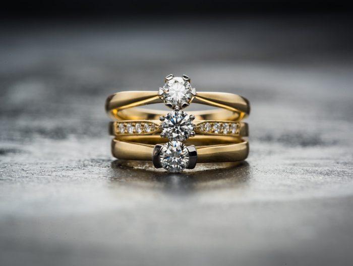 Three yellow gold engagement rings centered with diamonds stacked on a grey table.