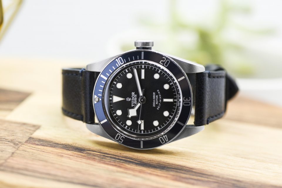 Pre-owned men’s Tudor in stainless steel with a black leather strap on a wooden table.