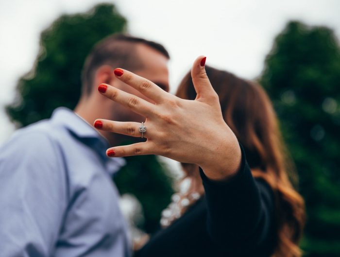 Woman’s hand featuring diamond engagement ring with couple kissing in background.