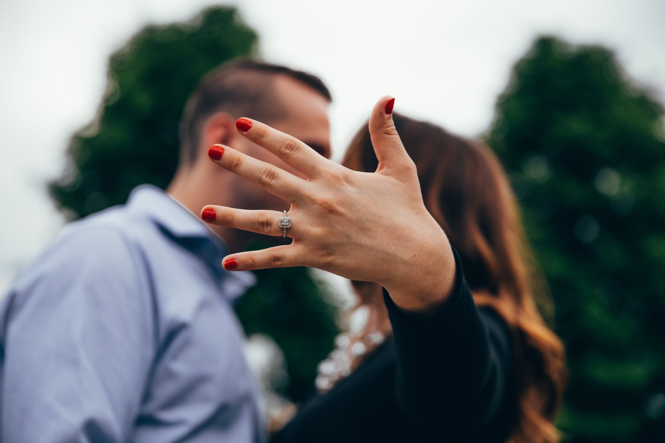 Woman’s hand featuring diamond engagement ring with couple kissing in background.