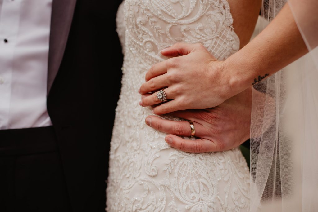 Bride and groom’s hands on bride’s waist featuring her diamond engagement ring and
their matching wedding bands.