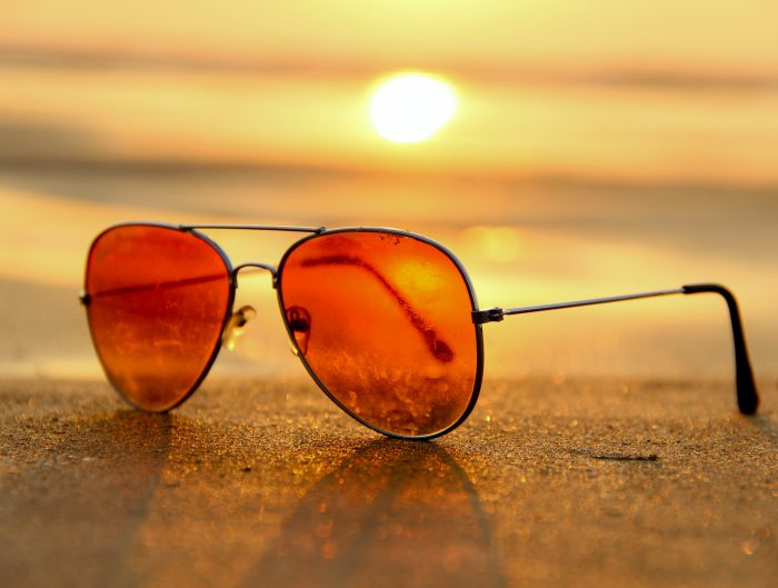 Rose colored aviator sunglasses in the sand with a beach sunset background.