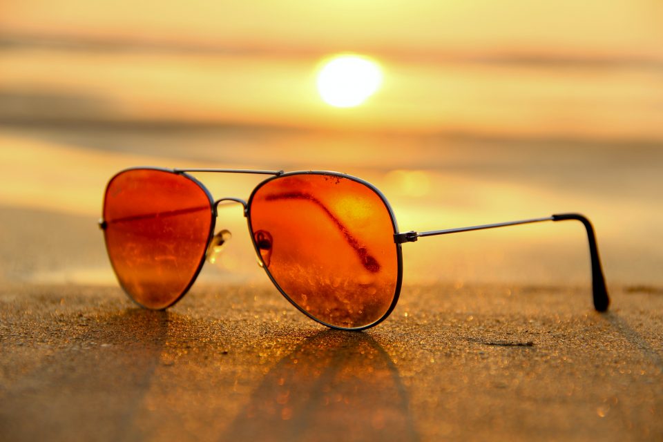 Rose colored aviator sunglasses in the sand with a beach sunset background.