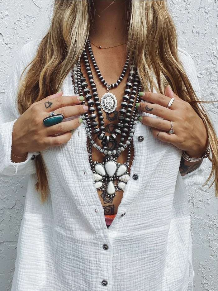 Torso portrait of woman against white wall wearing various stacked statement necklaces and rings.