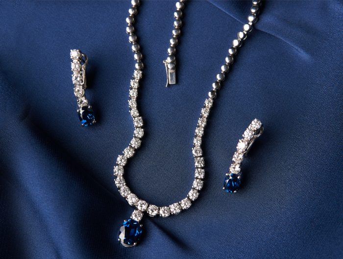 White gold necklace and earrings both set with diamonds and blue sapphires on a blue fabric background.