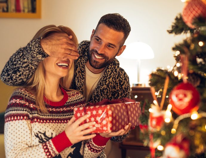 Man covering woman's eyes and giving her a gift in front of Christmas tree.