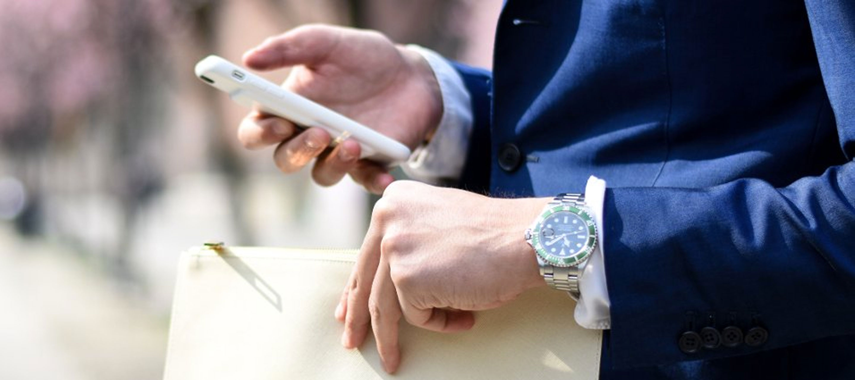 Close-up of a man in a blue suit on a street holding a phone and small clutch while
wearing a Rolex on left wrist.