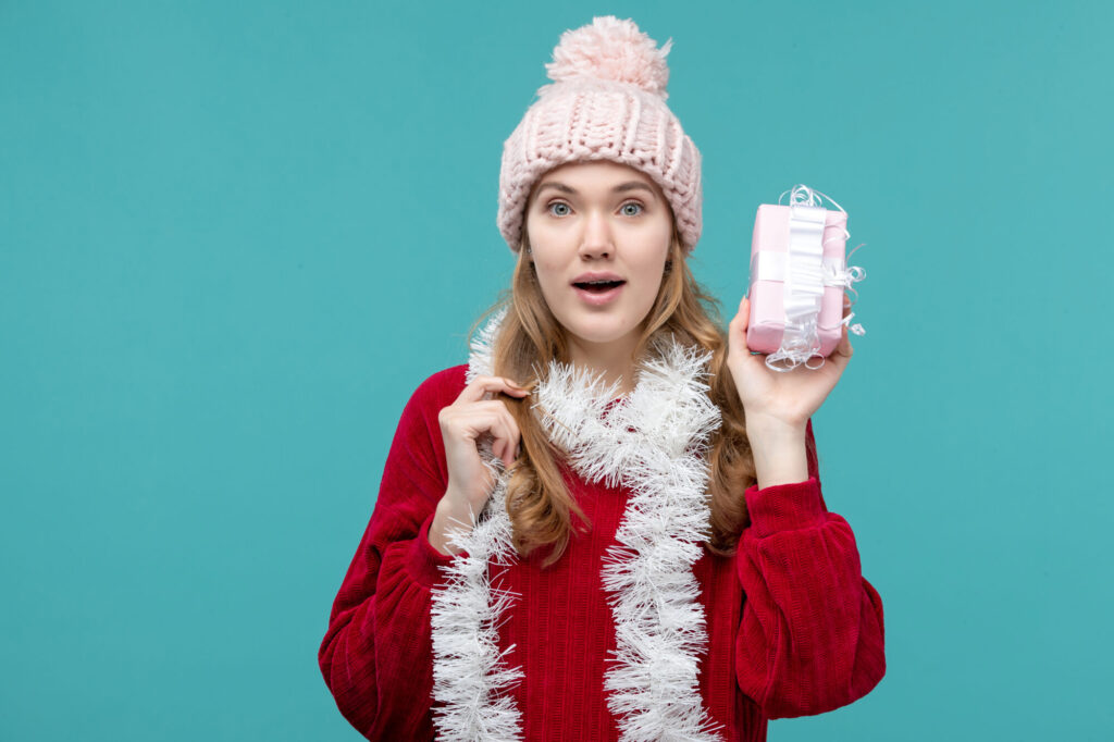 A photo of a woman in her 20s-30s wearing white Christmas tinsel as a scarf and a pink cable knit winter hat and red Christmas sweater, holding up a gift appearing to be the size of a necklace or bracelet in a gift box, wrapped in pastel pink paper and a cute white ribbon. Her expression looks to be that of a sister who is asking her sibling or family member what is in the box, or hoping that the present is something nice that she will like. 