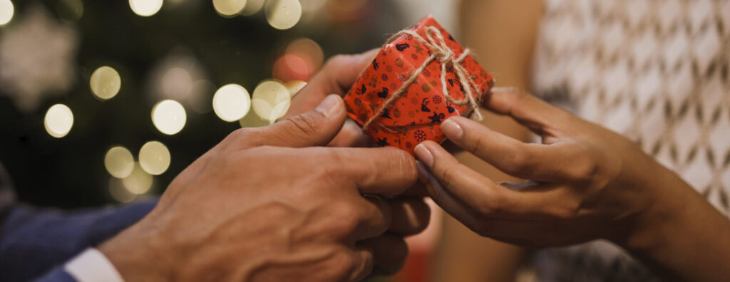 A closely shot photograph of a man handing a woman a small jewelry box wrapped in Christmas wrapping paper in front of holiday lights. 