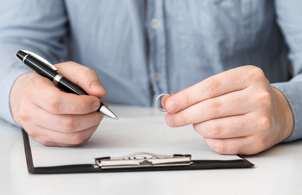 A close-up photograph of a person's hands, one holding a pen poised over a clipboard with a document, and the other holding a silver wedding band, suggesting a scene of signing legal papers.