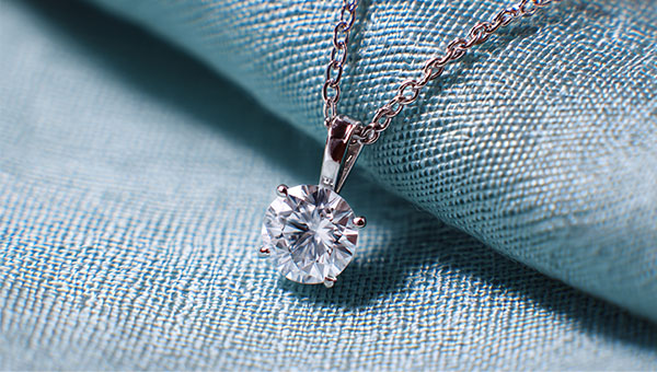 an image of a lab-grown diamond set in a white gold dainty pendant and attached to a white gold necklace. The diamond necklace rests on blue fabric.