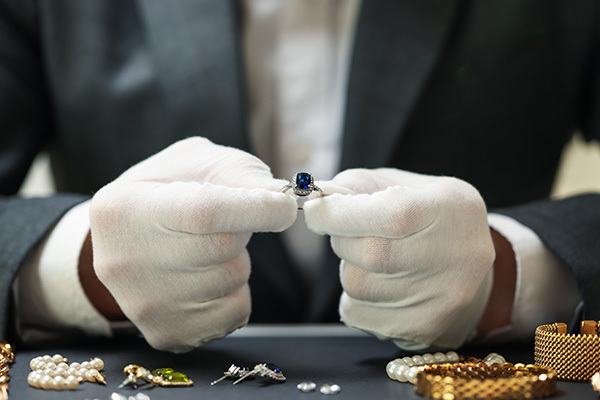 a close up image of a jeweler wearing white gloves holding a sapphire ring. various loose jewelry pieces is seen on the table next to the jeweler